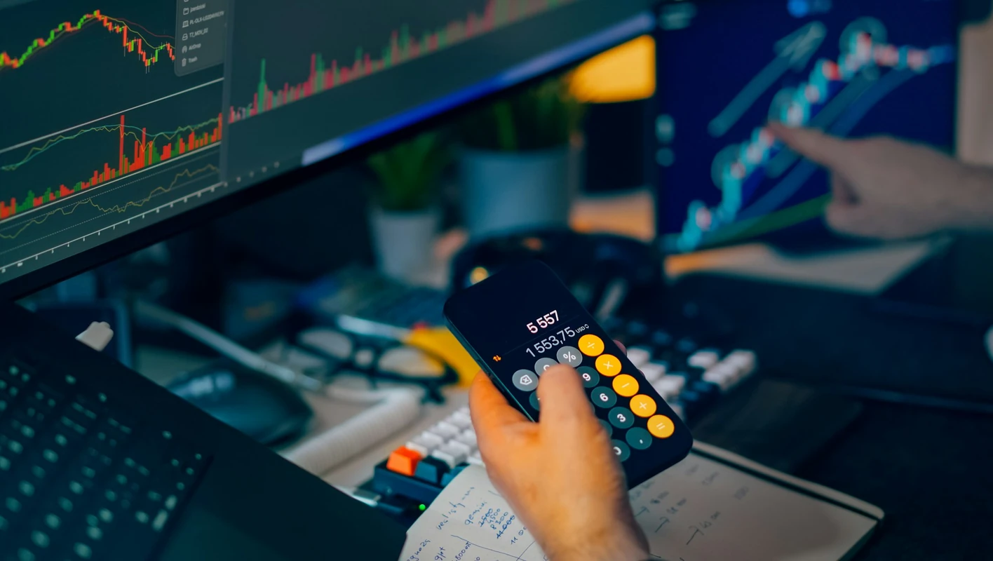 Profissional revisando cálculos em uma calculadora de celular diante de monitores com gráficos financeiros.